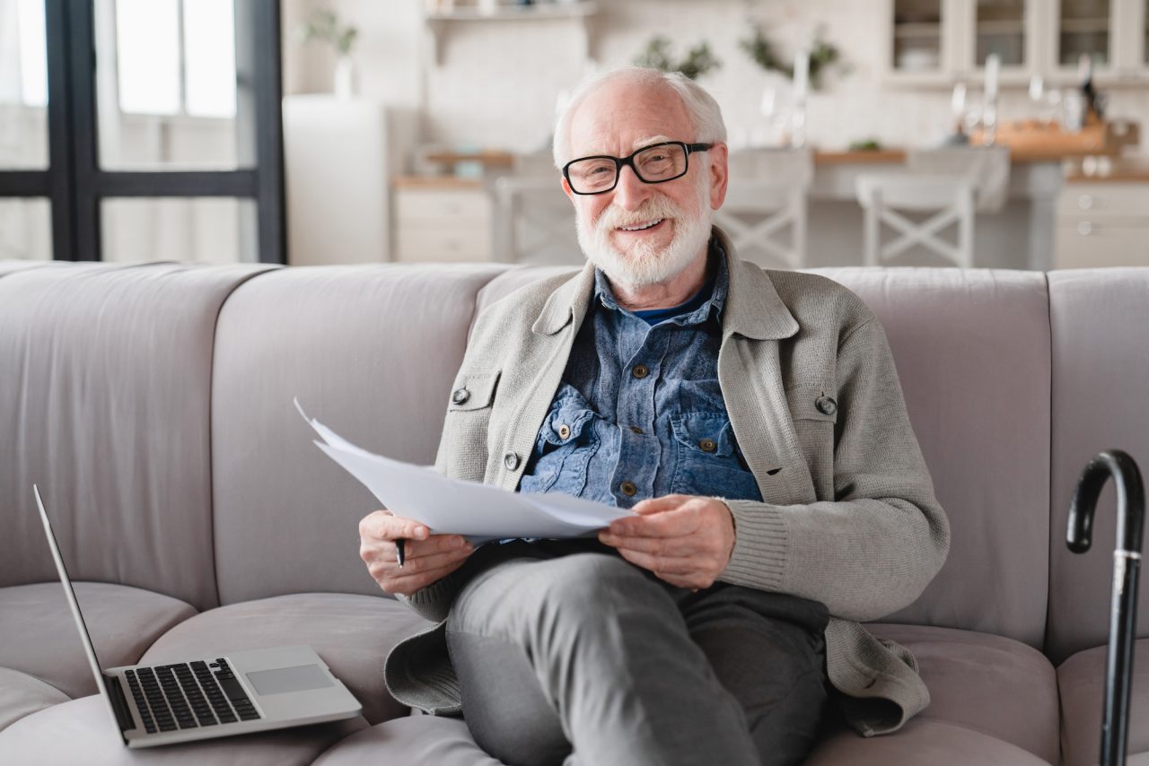 grandfather looking at camera checking pension documents loan e banking paperwork with laptop